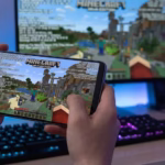 A person playing Minecraft Java Edition on an Android smartphone using PojavLauncher, with game debug information visible on the screen and a PC gaming setup in the background.